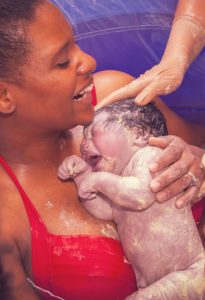 A happy African American mother holding her brand new baby girl after delivering her in a birthing pool at home.  The baby is covered in vernix and the mom is laughing.