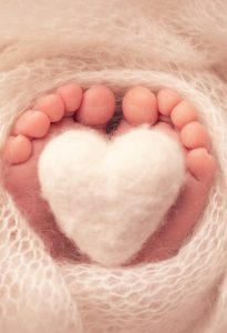 The tiny foot of a newborn baby. Soft feet of a new born in a white wool blanket. Close up of toes, heels and feet of a newborn. Knitted white heart in the legs of a baby. Macro photography.