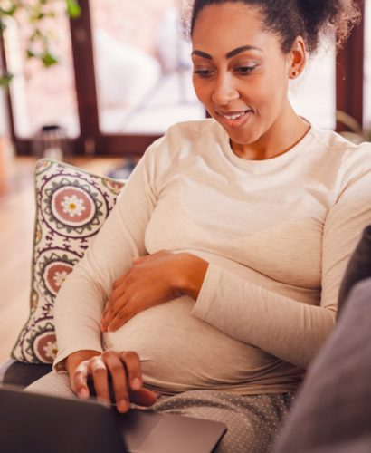 Adult beautiful pregnant smiling african woman working with laptop and holding her belly while sitting on sofa at home