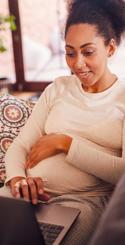 Adult beautiful pregnant smiling african woman working with laptop and holding her belly while sitting on sofa at home